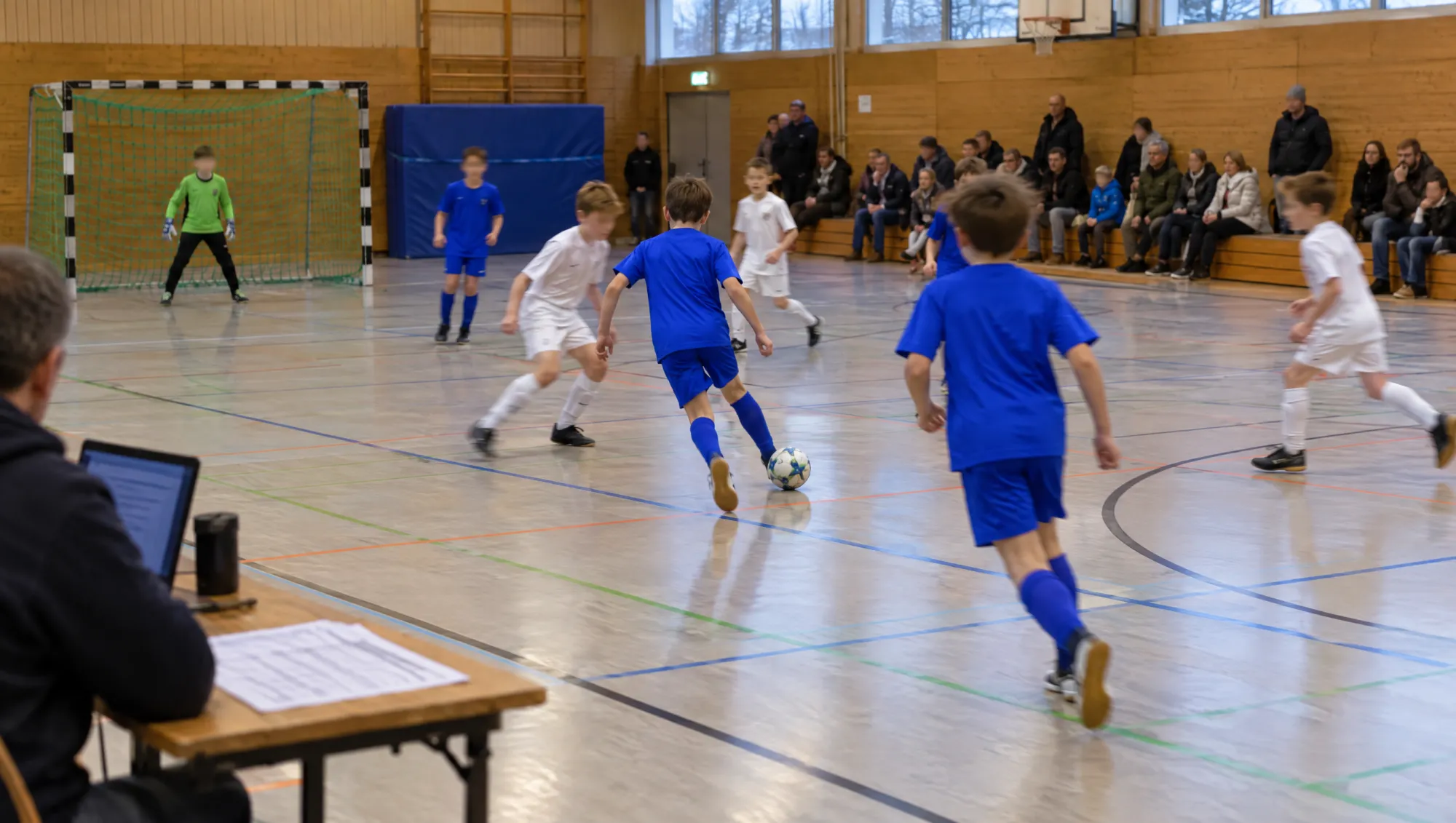Hallenturnier im Jugendfußball: Kinder in blauen und weißen Trikots spielen, vorne der Tisch der Turnierleitung mit ausgedrucktem Spielplan